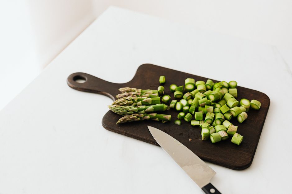 From above of chopped fresh asparagus near knife placed on dark cutting board composed on white table in kitchen